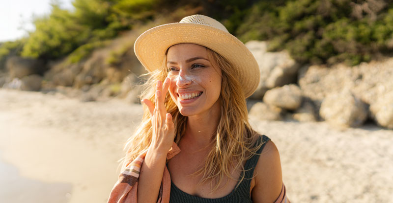 Eine lächelnde Frau mit Sonnenhut steht an einem Sandstrand und schützt ihre Augen mit der Hand vor der Sonne. Im Hintergrund plätschern sanft Wellen ans Ufer.