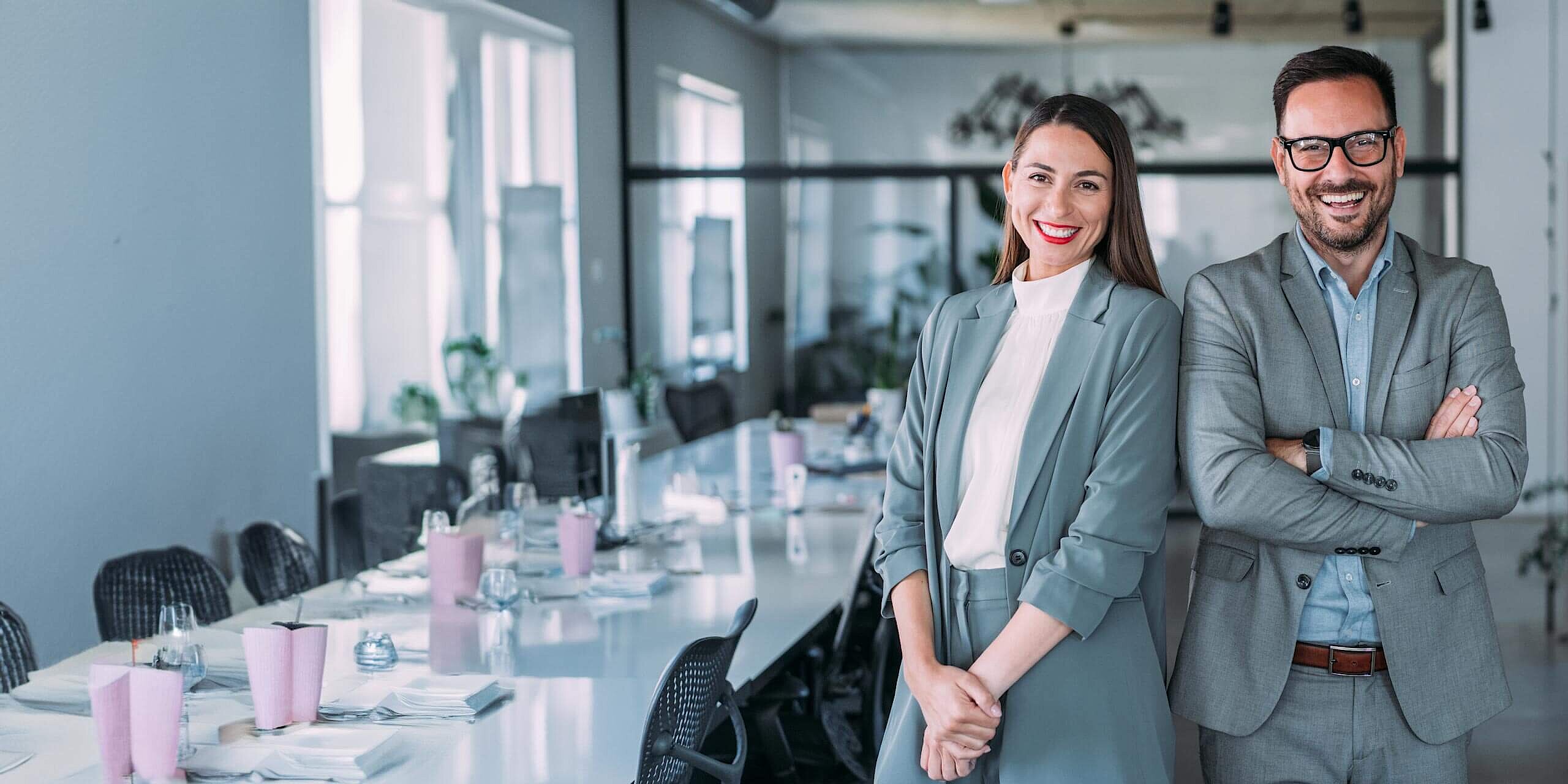 Ein Mann und eine Frau in grauen Business-Outfits stehen lachend in einem Meeting-Raum