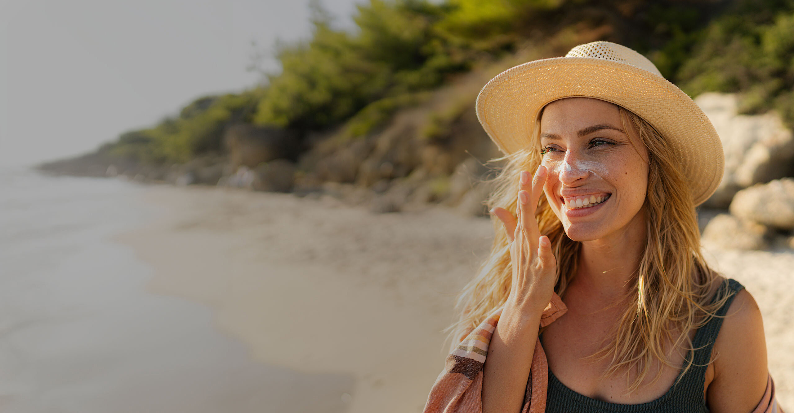 Eine lächelnde Frau mit Sonnenhut steht an einem Sandstrand und schützt ihre Augen mit der Hand vor der Sonne. Im Hintergrund plätschern sanft Wellen ans Ufer.